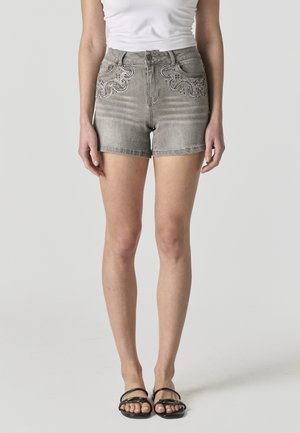 Mujer con shorts de mezclilla gris con diseños de paisley de pedrería en los bolsillos delanteros, camiseta blanca sin mangas y sandalias negras.
