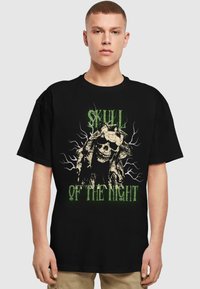 Sort t-shirt med et kranie-design og grøn tekst, der står "SKULL OF THE NIGHT", omgivet af intrikate grenmønstre.