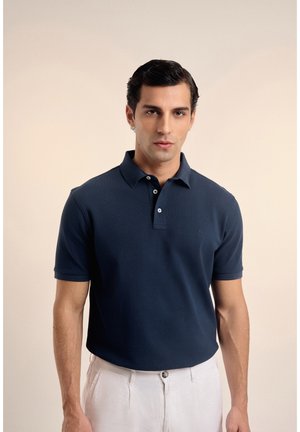 Hombre de cabello oscuro con polo azul marino y pantalones blancos, de pie frente a un fondo beige liso.