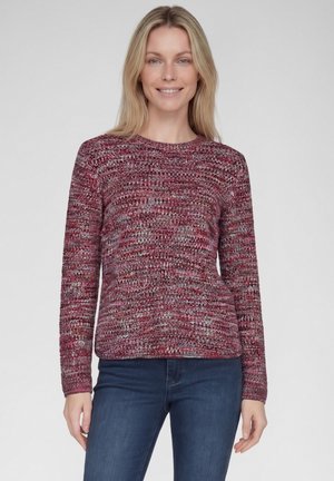 Woman with blonde hair wearing a maroon and gray knit sweater and blue jeans, standing against a plain light background.
