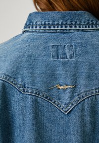 Stražnja strana traper jakne s oštrim jaramom, plava tkanina s izblijedjelom teksturom i izvezenim logotipom "Pepe Jeans" u zlatnoj boji.