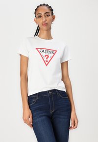 T-shirt en coton blanc avec un logo triangulaire rouge central portant le texte "GUESS®". Le modèle porte un jean bleu foncé, mettant en valeur un design ajusté.