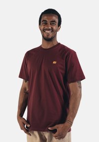 Bordeauxrotes Baumwoll-T-Shirt mit kurzen Ärmeln und rundem Ausschnitt, das auf der linken Brust ein kleines gesticktes Elefantenlogo in Orange zeigt.