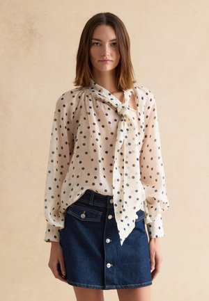 Mujer joven con una blusa de gasa blanca con lunares, cuello atado y mangas largas, combinada con una falda de mezclilla oscura abotonada.