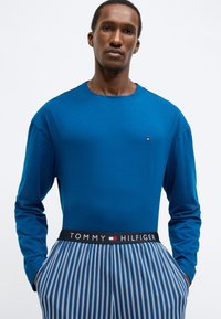 Μπλε πουκάμισο μακρυμάνικο με στρογγυλή λαιμόκοψη, συνδυασμένο με ριγέ παντελόνι πιτζάμας με μαύρη ζώνη στη μέση που φέρει το λογότυπο "TOMMY HILFIGER".