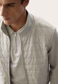 Giacca bomber grigia chiara con texture trapuntata sul davanti, colletto a coste e chiusura a zip quarti, abbinata a una polo coordinata.