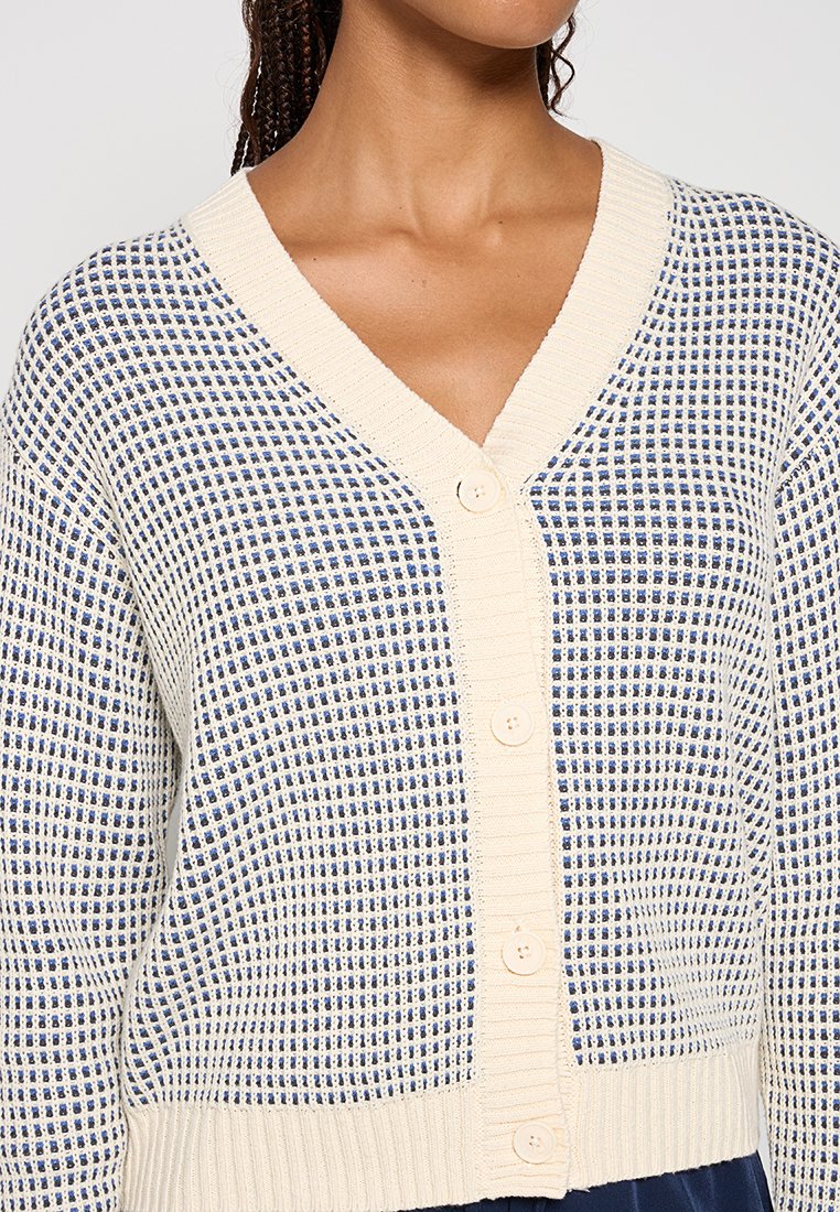 Sweter kardigan z dekoltem w serek w odcieniu kremowym, z niebieskim wzorem w kratę, ribbowanym dołem i mankietami oraz sześcioma przyciskami z przodu do zapięcia.