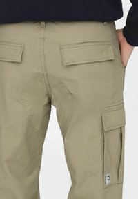 Beiget väriset cargo-housut, joissa on teksturoitu kangas, kaksi takataskua läpällä ja vahvistetut polvitaskut. Sivutaskussa on logoyksityiskohta.