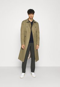 Olive green trench coat with double-breasted design, large buttons, and a smooth texture; paired with dark trousers and white sneakers.