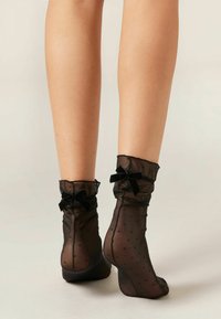 Sorte netankelsokker med et prikket mønster, der har flæsekanter og små sorte velour-sløjfer ved mansjetterne.