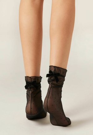 Sorte netankelsokker med et prikket mønster, der har flæsekanter og små sorte velour-sløjfer ved mansjetterne.