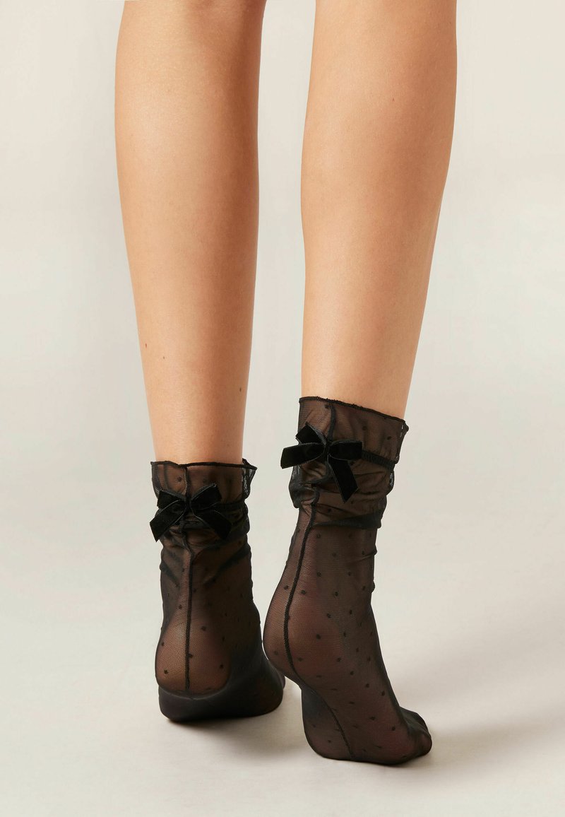 Sorte netankelsokker med et prikket mønster, der har flæsekanter og små sorte velour-sløjfer ved mansjetterne.