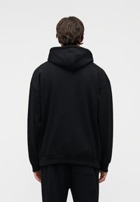 Μαύρο φούτερ με κουκούλα και χαλαρή εφαρμογή, με υφή στο ύφασμα και ριμπ τελειώματα. Ο σχεδιασμός έχει μια απλή, μινιμαλιστική αισθητική.