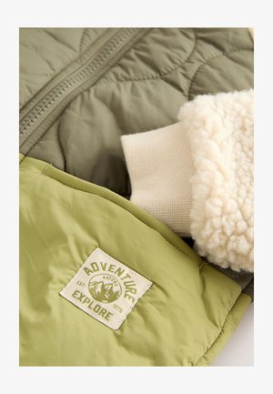 Gewatteerde groene jas met een gestructureerde beige fleece kraag, voorzien van een olijfgroene patch met de tekst "ADVENTURE EXPLORE" en een ritssluiting.