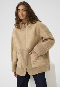 Naine, kes kannab helepruunist tõmblukuga sherpa-jakki suurte eesmiste taskutega ja tumedaid pükse, seisab tavalise valge taustaga.