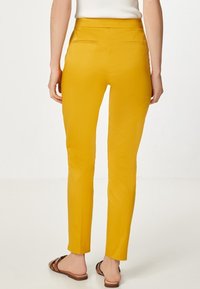 Pantaloni slim fit giallo senape indossati da una persona in piedi, abbinati a sandali flat marroni e a una maglietta bianca senza maniche.