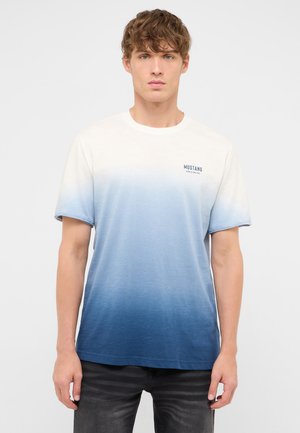 Junger Mann mit kurzem gewelltem Haar trägt ein kurzärmliges weiß-blau verlaufendes T-Shirt und dunkle Jeans, steht vor einem schlichten hellen Hintergrund.