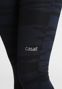 Černé legíny s tmavým maskáčovým vzorem, které mají vyšívané logo "casall" po straně a hladkou, elastickou texturu.