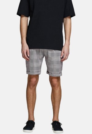 Man in een zwarte oversized t-shirt, grijze geruite shorts met omgeslagen hemden en zwarte sneakers met witte zolen, staand tegen een effen achtergrond.