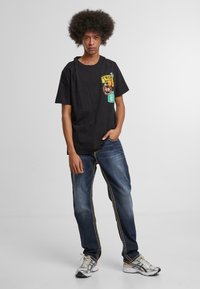 Camiseta negra con estampado gráfico y acentos de color combinada con jeans azules con costuras y bolsillos amarillos. Zapatillas deportivas grises.