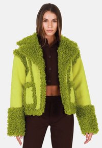 Vihreä fleece-takki, jossa on teksturoidut kiharat reunat, hupullinen, etutaskut ja lyhyt muotoilu. Yhdistetty ruskean paidan ja housujen kanssa.