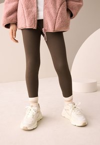 Barna bordázott leggings, krémszínű sportcipővel és csíkos zoknival párosítva. A modell egy rózsaszín polárdzsekit visel cipzáras lezárással.