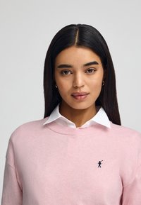 Maglione rosa chiaro con collo rotondo e maniche lunghe, indossato sopra una camicia bianca con colletto, con un piccolo logo ricamato in nero.