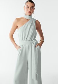 Viena pleca kombinezons gaiši zaļā krāsā ar strukturētu audumu, ar savilktu jostasvietu un sānu kabatām. Elegants, minimālistisks dizains.