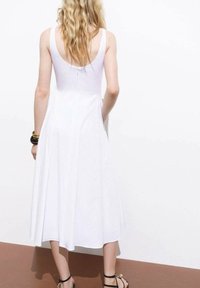 Robe blanche sans manches avec une silhouette évasée, en tissu lisse et avec un dos nu. Le modèle porte des sandales noires à lanières et des bracelets métalliques.