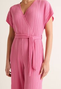 Rosa plisserad jumpsuit med omslagdesign, korta ärmar och ett matchande bälte. Texturen är slät med vertikala ränder.