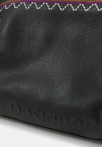 Schwarze Lederhandtasche mit strukturierter Oberfläche, ausgestattet mit weißen und pinken Zickzackstichen sowie geprägtem "Desigual"-Logo-Detail.