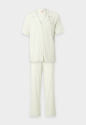 Joven mujer de pie descalza con una camisa blanca holgada de manga corta abotonada y pantalones anchos blancos a juego sobre un fondo liso.