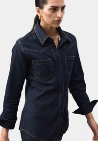 Mørk denimskjorte med spids krave, to brystlommer, orange syninger og lange ærmer med oprevet mansjetter.