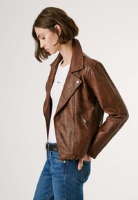 Bruine leren motorjack met een asymmetrische rits, drukknopenkraag en een gestructureerde afwerking. Gedragen over een wit shirt met blauwe jeans.