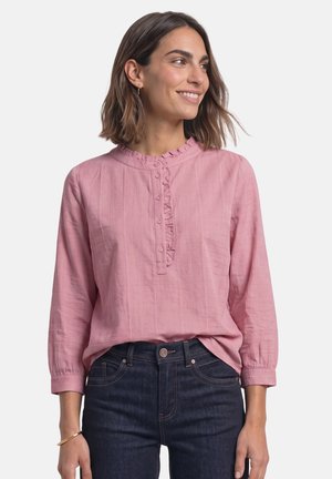 Frau lächelt und schaut nach rechts, trägt eine langärmlige rosa Bluse mit Rüschen und dunkelblaue Jeans vor einem einfarbigen hellen Hintergrund.
