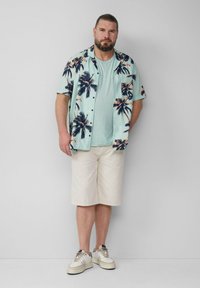 Camicia azzurra a maniche corte con stampa di palme, abbinata a una t-shirt verde chiaro e pantaloni beige; indossa sneakers bianche con dettagli.
