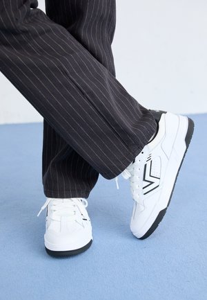 Sneakers bianchi con accenti neri e un design geometrico. Posizionati su una superficie blu, abbinati a pantaloni a righe sottili scure.