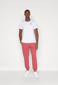 Balts Nike T-krekls ar melnu logotipu, savienots ar rozā sporta biksēm un baltām kedām. Vienkāršs dizains, pieguļošs siluets, gluda auduma tekstūra.
