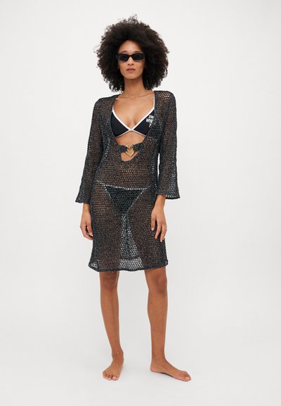 Cubierta de malla negra con mangas largas, frente abierto y detalle en forma de corazón. Llevada sobre un bikini negro. Textura suave y ajuste holgado.