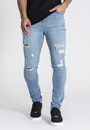 Light blue denim jeans featuring distressed details with rips and paint splatter, paired with black sneakers. Fitted design, tapered legs.