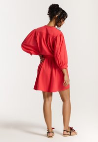Rotes, kurzes Kleid mit gerafften Ärmeln und taillierter Taille, aus leichtem Stoff gefertigt. Getragen mit flachen schwarzen Sandalen mit dekorativen Akzenten.