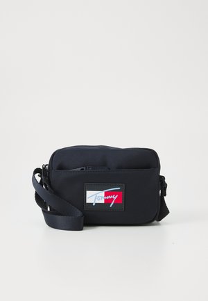 Jūras zila crossbody soma, kas izgatavota no izturīga auduma, ar rāvējslēdzēja aizdari, regulējamu siksnu un logo plāksni ar sarkaniem, baltiem un ziliem akcentiem.