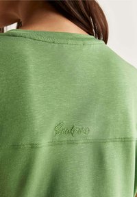 Femme portant un haut vert clair avec un tissu texturé et un petit logo brodé "Seadpods" sous la couture du décolleté, mettant en valeur le haut du dos et le cou.