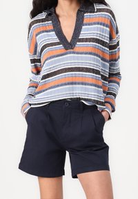 Randig stickad topp med blå, orange och svarta linjer; har en V-ringning och krage. Matchas med mörk marinblå shorts.