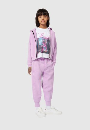 Fille debout portant un hoodie violet clair et un jogging, un t-shirt blanc avec un graphique et du texte rose, des chaussettes blanches et des baskets blanches sur un fond gris.