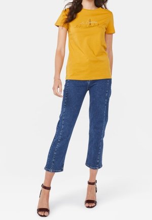 Gelbes Baumwoll-T-Shirt mit kurzen Ärmeln, das ein grafisches Design auf der Vorderseite zeigt. Kombiniert mit blauen, taillierten Jeans und braunen, hohen Sandalen.