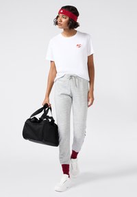 T-shirt bianco a maniche corte con un piccolo logo a forma di cuore, abbinato a pantaloni della tuta grigi chiari, calzini rossi e sneakers bianche. Borsa a tracolla nera inclusa.