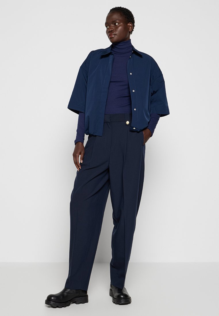 Femme portant un col roulé bleu marine, une veste à manches courtes, un pantalon large taille haute et des bottes noires chunky devant un mur blanc.