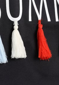 Černá tkanina s výrazným bílým textem "LÉTO", s třemi ozdobnými střapci: světle modrým, bílým a červeným, každý s texturovaným povrchem.