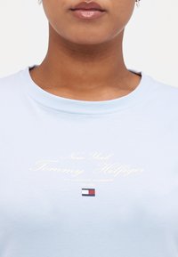 T-shirt en coton bleu clair avec un texte brodé blanc : « New York Tommy Hilfiger », complété par un petit logo bleu marine, rouge et blanc en dessous.